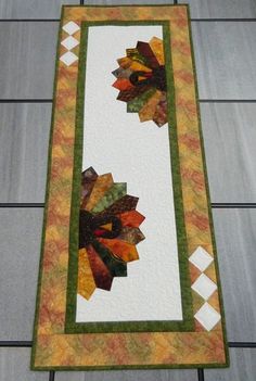 a quilted table runner with an orange and green flower on the center, along with two white squares