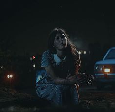 a woman sitting on the ground in front of a car at night with her eyes closed