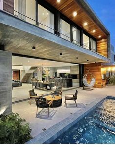 an outdoor dining area with chairs and tables next to a pool at night, in front of a modern home