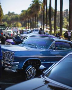 an old blue car parked in a parking lot next to other cars and palm trees