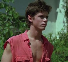 a shirtless young man standing in front of a house with trees and bushes behind him