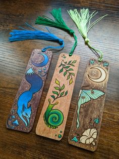 three wooden bookmarks with different designs on them sitting on a table next to some tassels