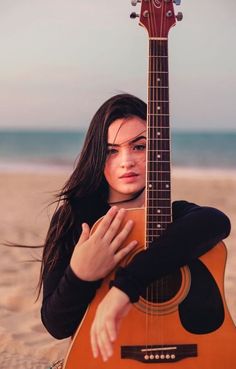 a woman sitting on the beach with her guitar in front of her face and looking at the camera