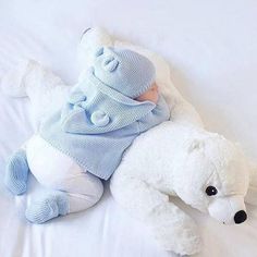 a white teddy bear laying on top of a bed next to a baby wearing a blue sweater