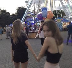 two young women holding hands in front of a carnival ride at night with people walking around