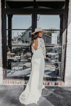 a woman standing in front of a window wearing a white dress and hat with her back to the camera