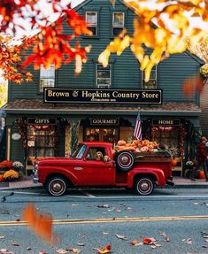 an old red truck parked in front of a store with autumn leaves on the ground