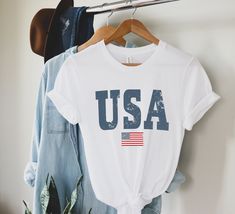 Celebrate your patriotism with our oversized varsity letter USA tee -- our graphic has been distressed to give it a vintage vibe. Perfect for you entire family! Youth sizes here: https://www.etsy.com/listing/1024874437/kids-usa-shirt-retro-usa-t-shirt-youth?ref=shop_home_active_2&pro=1&frs=1 Looking for more 4th of July gear? Check these out: https://www.etsy.com/shop/TheGraphicPeach?ref=seller-platform-mcnav&section_id=33390667 Our shop uses direct-to-garment printing to make our pr Americana Cotton T-shirt With Letter Print, 4th Of July Streetwear T-shirt With Letter Print, White Americana Letter Print T-shirt, 4th Of July Cotton T-shirt With Text Print, White T-shirt With American Flag Print, Casual Letter Print T-shirt For Independence Day, American Style White Tops With Letter Print, White American Style Tops With Letter Print, White Americana T-shirt With American Flag