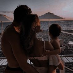 a woman holding a baby on the beach at dusk with umbrellas in the background