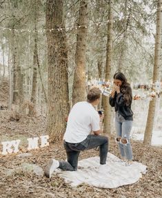 a man kneeling down next to a woman on a blanket in the woods with string lights