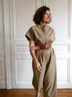 a woman standing in front of a white wall wearing a brown checkered jumpsuit