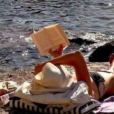 a woman reading a book while laying on the ground next to some water and rocks