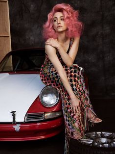 a woman with pink hair sitting next to a red car and posing for the camera