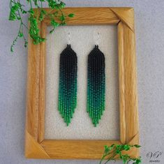 two green and black beaded earrings in a wooden frame next to a plant on the floor