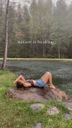 a woman laying on top of a rock next to a body of water with trees in the background