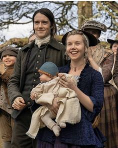 a man and woman holding a baby in front of other people wearing period clothing,