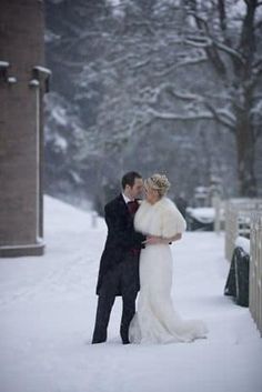 a bride and groom standing in the snow near a fence with their arms around each other