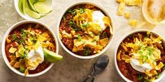 three white bowls filled with chili, sour cream and tortilla chips on top of a table