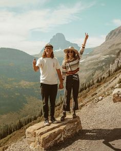 two people standing on top of a mountain holding hands and looking into the distance with mountains in the background