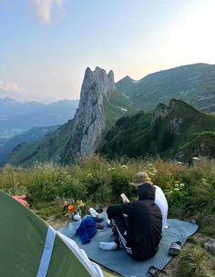 a man sitting on top of a green field next to a tent in the mountains