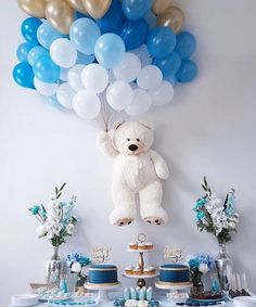 a white teddy bear sitting on top of a table next to blue and gold balloons