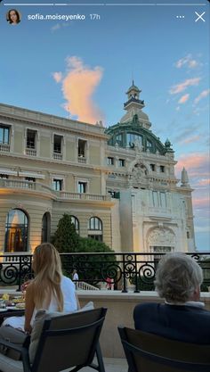 two people sitting on chairs in front of a large building with an ornate balcony and balconies