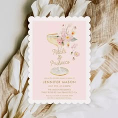 a pink and white wedding card with flowers on the bottom, in front of some feathers