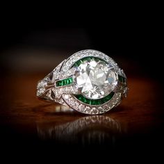 a diamond and emerald ring sitting on top of a wooden table in front of a black background