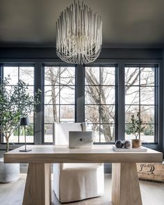 a desk with a laptop on it in front of two windows and a chandelier hanging from the ceiling