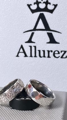 two silver wedding rings sitting on top of a white box with the word allurez written