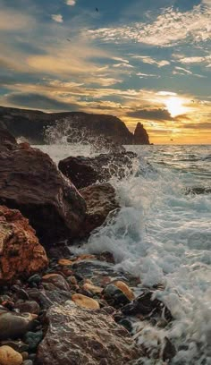 the sun is setting over the ocean with rocks and waves crashing on it's shore