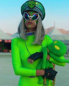 a woman in a green outfit holding a stuffed animal and wearing sunglasses with an alien hat on her head