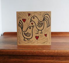 a card with two roosters on it sitting on top of a wooden table next to a wall