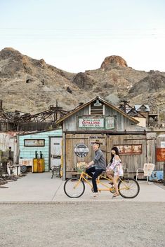 a man and woman are riding a bike in front of an old building with mountains in the background