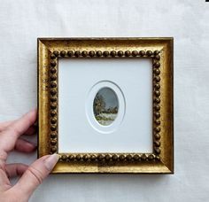 a person is holding up a painting in a gold frame on a white tablecloth