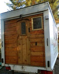 a small trailer with a wooden door and windows on the side, parked in front of some trees