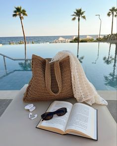 an open book sitting on top of a table next to a bag and sunglasses near a pool