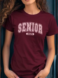 🌟 Stand Out in Your Senior Year with Our Vibrant Collegiate T-Shirt! 🌟 Get ready to celebrate your senior year with style and confidence with our eye-catching Senior Year T-Shirt in Cardinal Red! This design features the word "SENIOR" in large, blocky, and bold Collegiate letters, reminiscent of collegiate and varsity styles, perfect for making a statement. Below "SENIOR," the year "2025" is displayed in a similar bold font within an oval shape, adding a distinctive touch. The entire text and College Style Crew Neck T-shirt For College Events, Graphic Tee With Crew Neck For College Events, Text Print Crew Neck Top For College Events, Graphic Tee Crew Neck For Graduation, Collegiate Short Sleeve Tops For College Events, College Style T-shirt With Letter Print, Letter Design, School Events, Dynamic Design