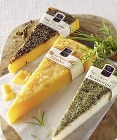 four different types of cheese on a wooden platter with herbs and seasoning around them