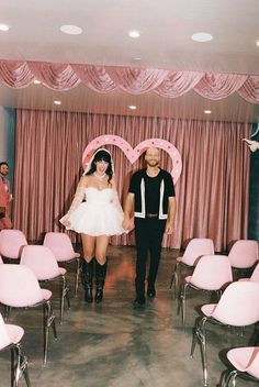 a man and woman standing in front of pink chairs with hearts on the wall behind them