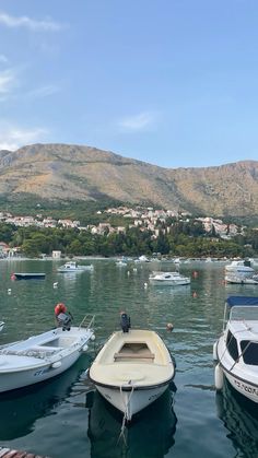 several small boats are docked in the water near some hills and buildings, with mountains in the background