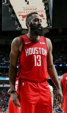 a man with a beard standing in front of a basketball court wearing red uniform and looking off to the side