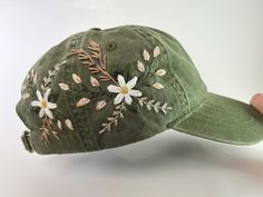 a green hat with white daisies and leaves embroidered on the front, held up by a person's hand