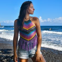 a beautiful young woman standing on top of a beach next to the ocean wearing a colorful crocheted halter top