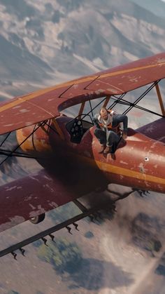 a man is sitting on the wing of an old biplane flying over a mountain range