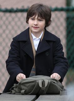 a young boy sitting on top of a bench next to a fence and holding onto a bag