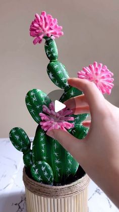 a hand holding a fake cactus plant with pink flowers in it's palm, on a white table