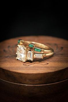 two gold rings with emeralds and diamonds on top of a wooden box in the dark