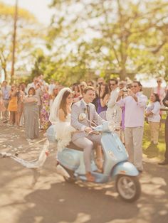 a bride and groom ride on a blue scooter in front of a group of people