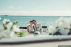 a bride and groom are sitting on a bench by the ocean with flowers in front of them